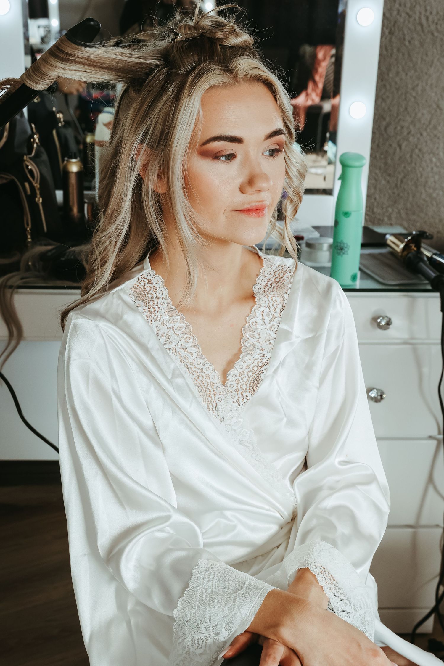 A person in a white satin and lace robe sits at a vanity while getting ready for a special occasion.