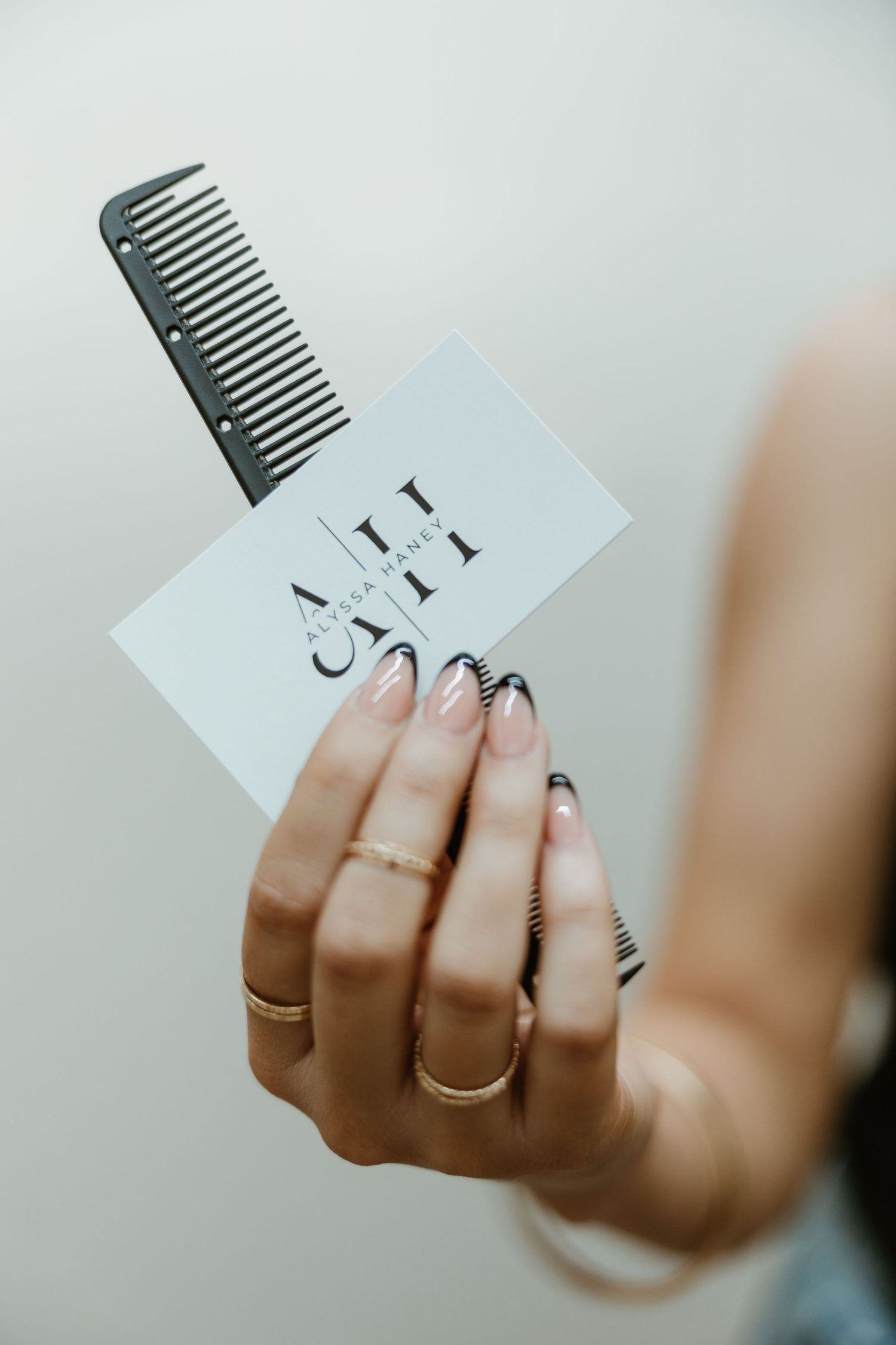 Manicured hand with multiple rings holding a white business card and gray comb against light background.