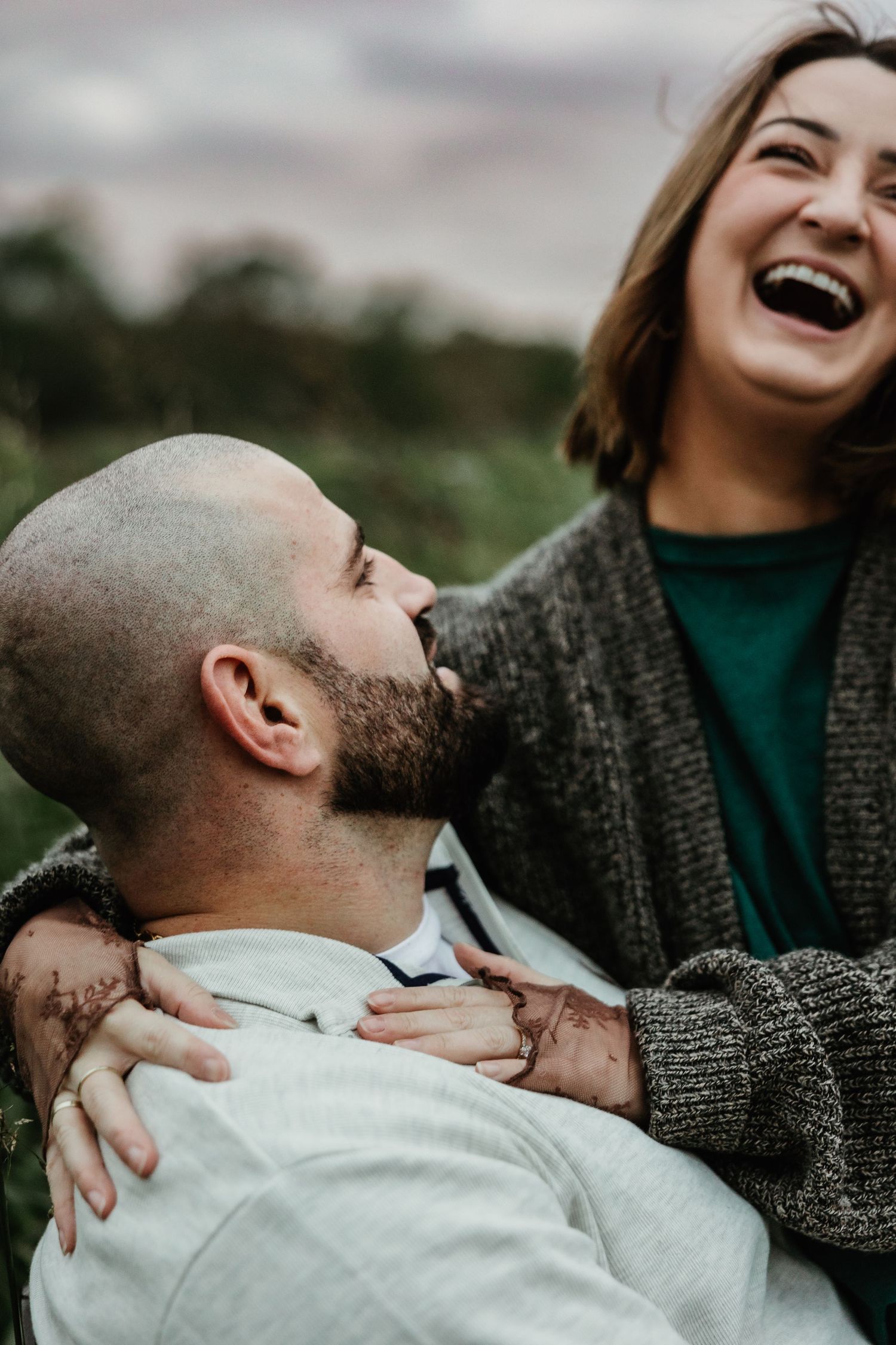 A couple share a joyful moment together outdoors, laughing while wearing casual sweaters on an overcast day.