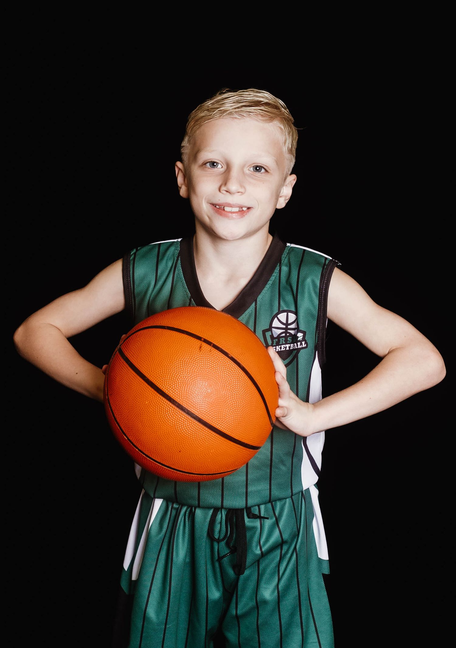 Young basketball player in green striped jersey poses with orange basketball against dark background.