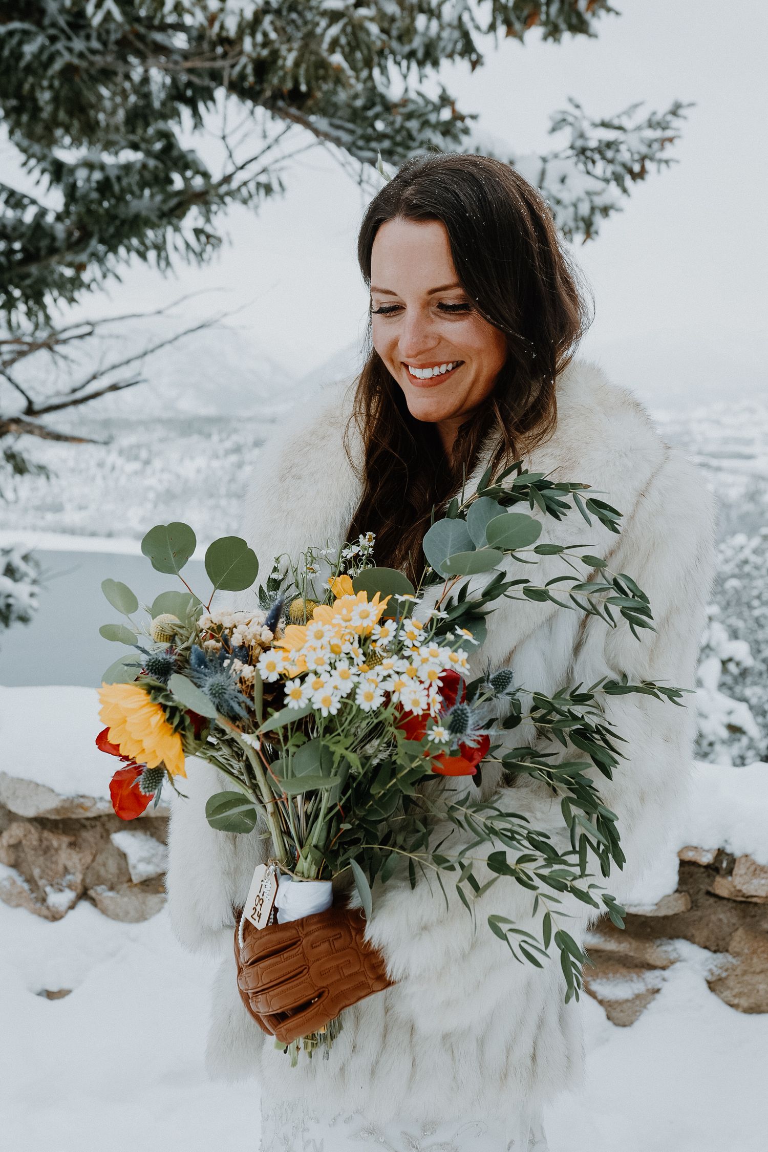 Winter bride in white fur coat holds colorful bouquet of yellow flowers and eucalyptus against snowy mountain backdrop.