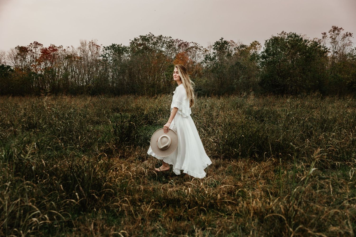Ethereal figure in flowing white dress walks through autumn field at dusk holding wide-brimmed hat.