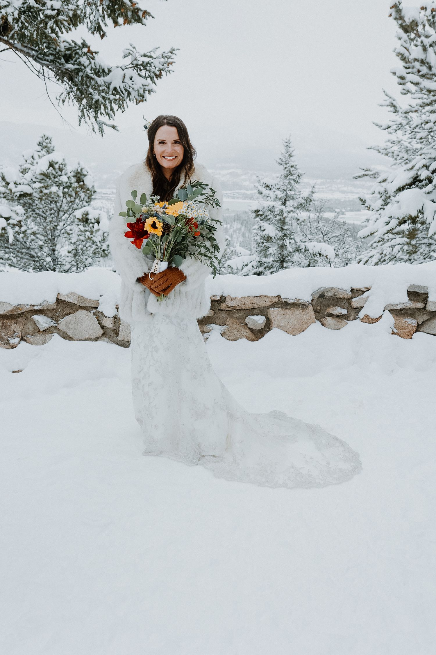 A bride in a white lace gown holds a colorful bouquet while standing in a snowy winter landscape with stone wall.