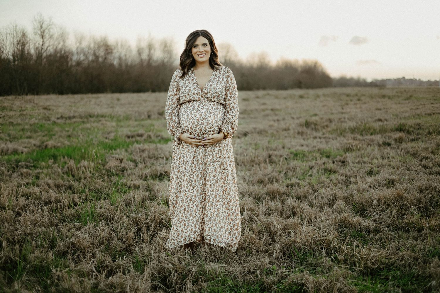 A person wearing a long floral dress stands in an open field at sunset during a maternity photoshoot.