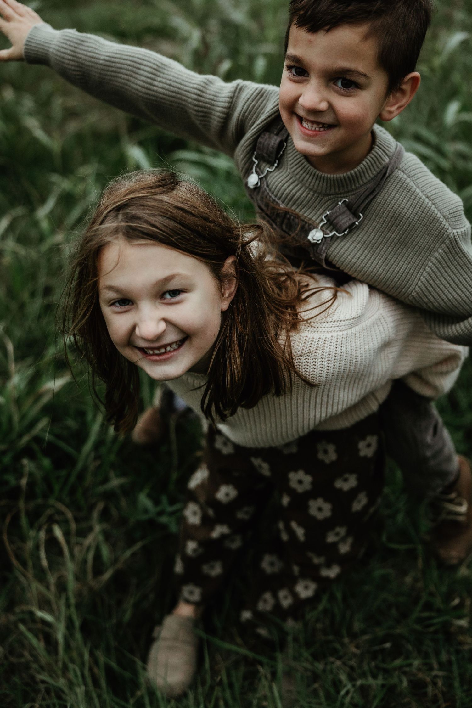 Two children in sweaters play and laugh together in tall green grass during an outdoor photo session.