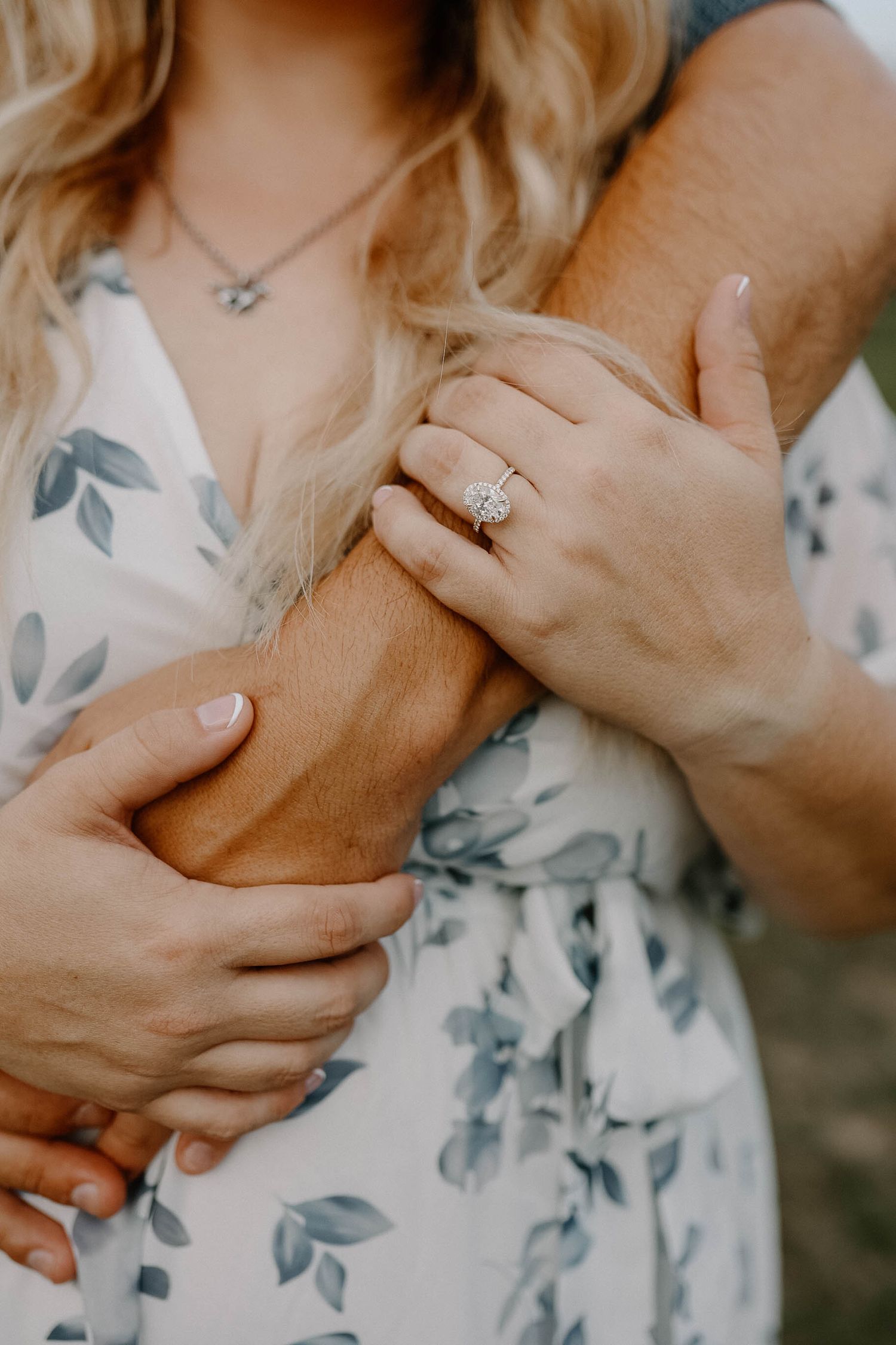 Close-up of hands wearing engagement ring and floral dress embracing arm in tender romantic pose against neutral background.