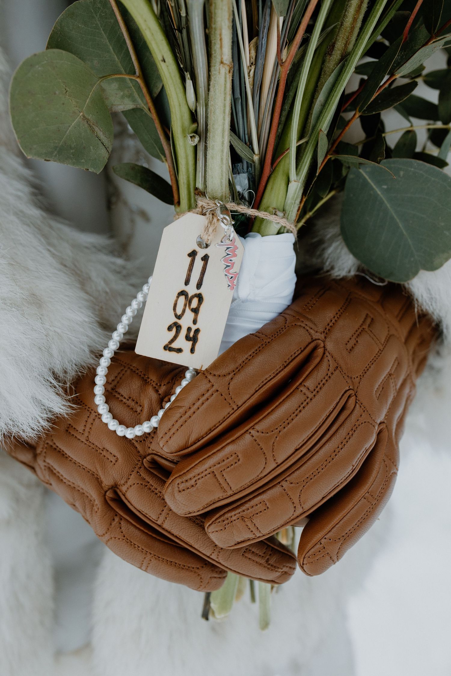 Brown leather baseball glove holds eucalyptus stems with a date tag and pearl bracelet in a styled wedding detail shot.