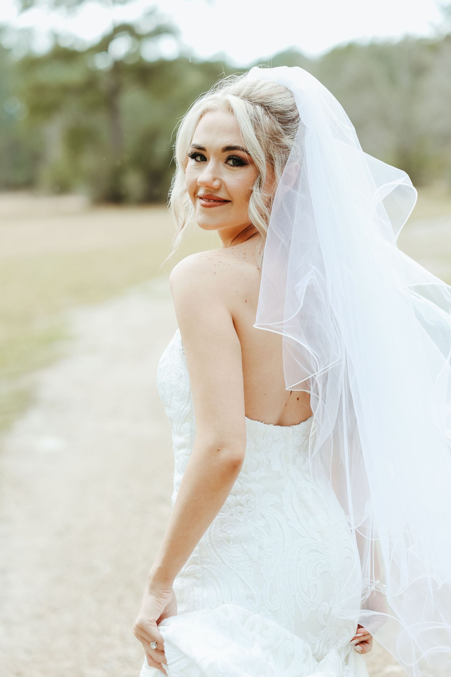 A bride in a white lace wedding dress and flowing veil looks over her shoulder while standing outdoors.