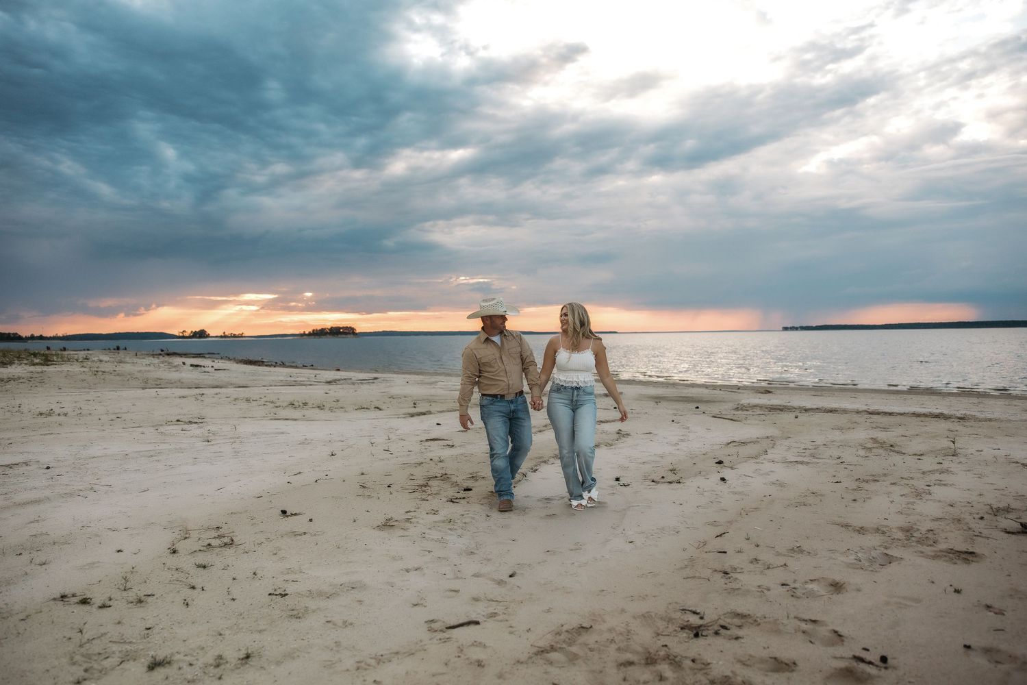 A couple walking hand in hand along a beach at sunset with dramatic cloudy skies overhead.