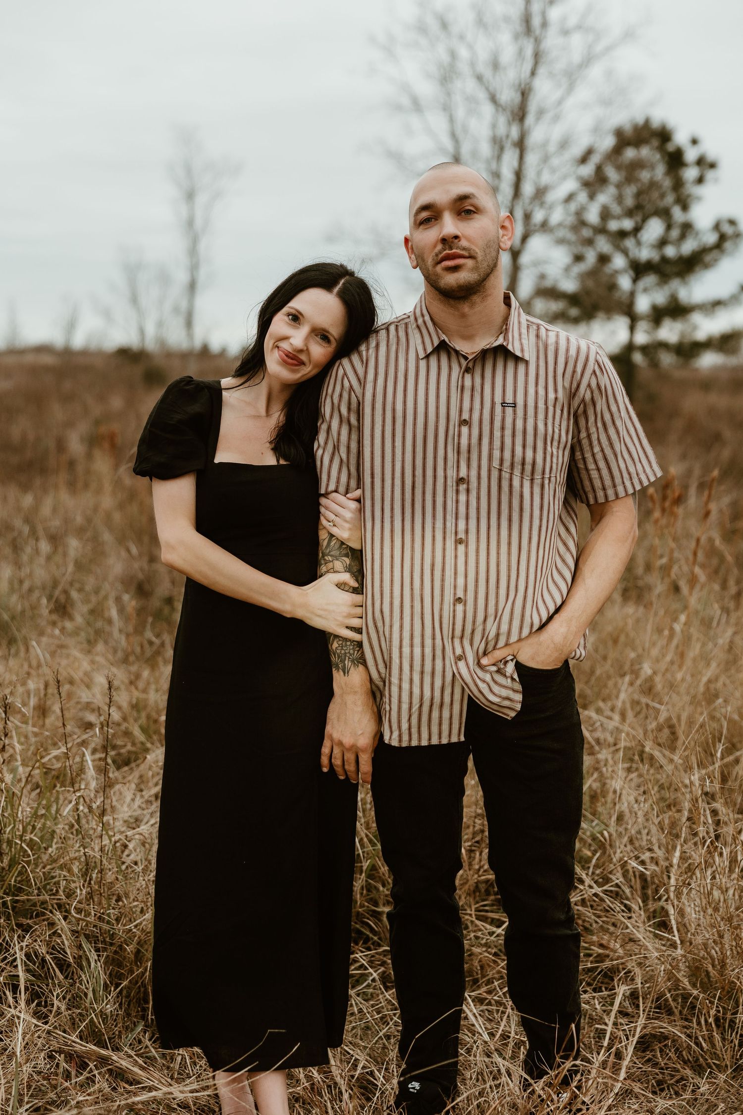 A couple poses together in a grassy field on an overcast day, her in black dress and him in striped shirt.