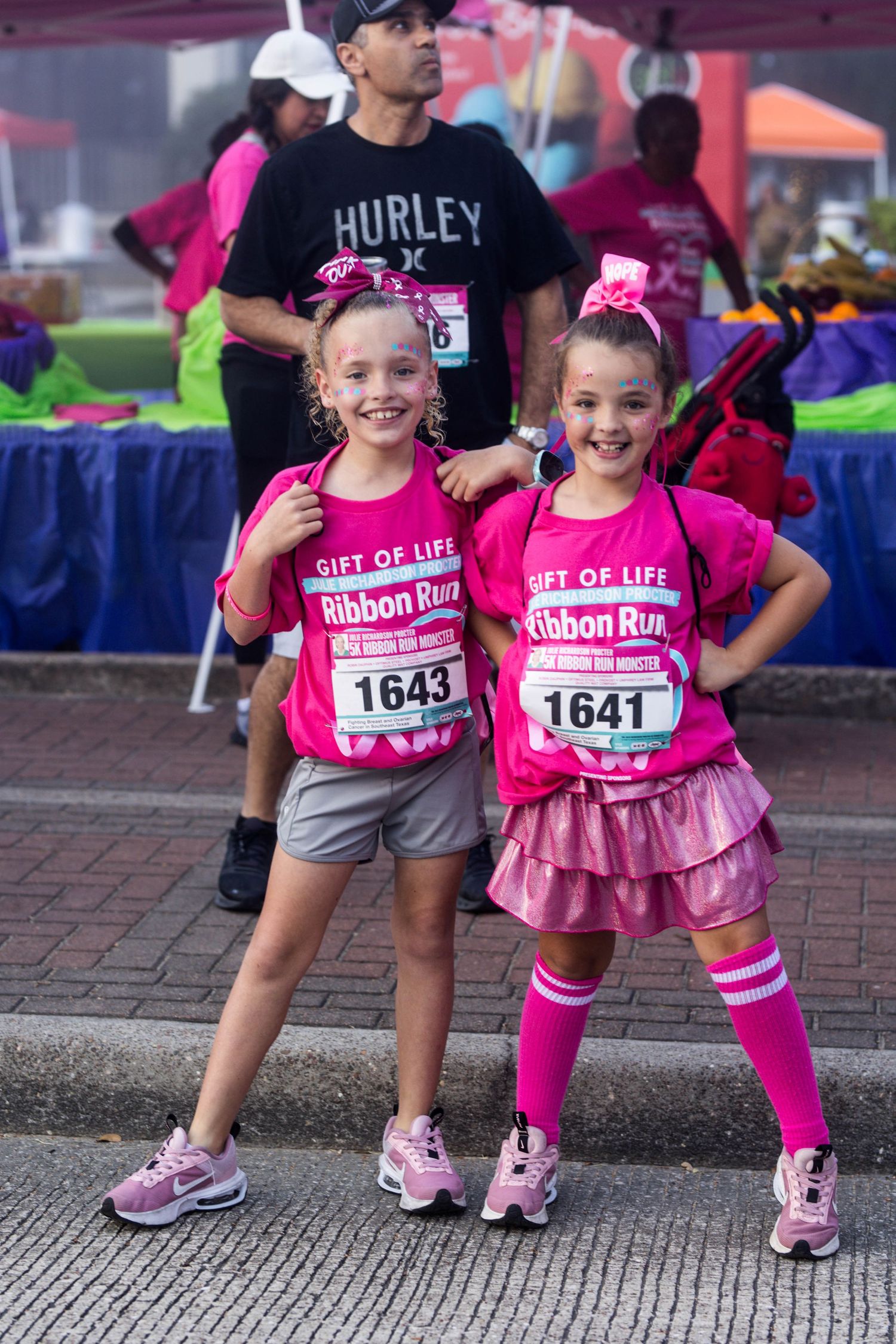 Two young race participants in pink attire pose together at a breast cancer awareness Ribbon Run event.