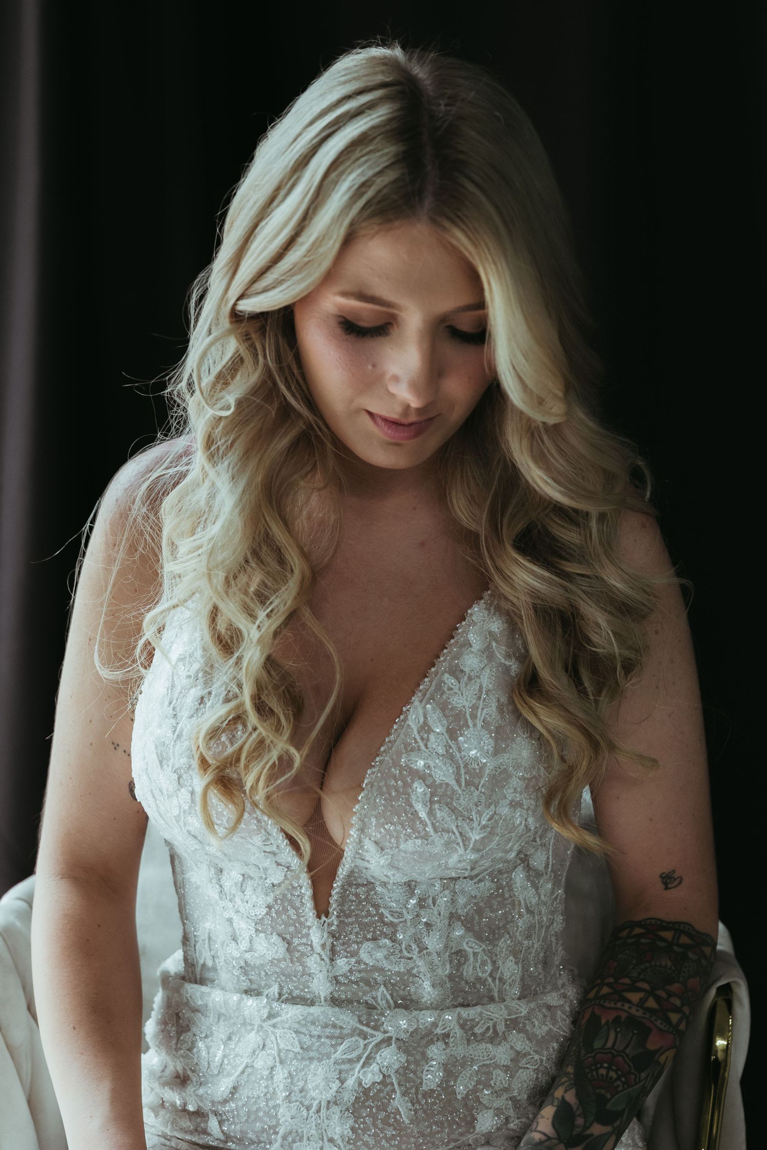 A bride wearing a delicate white lace wedding dress with long wavy blonde hair looks down in a moody portrait.
