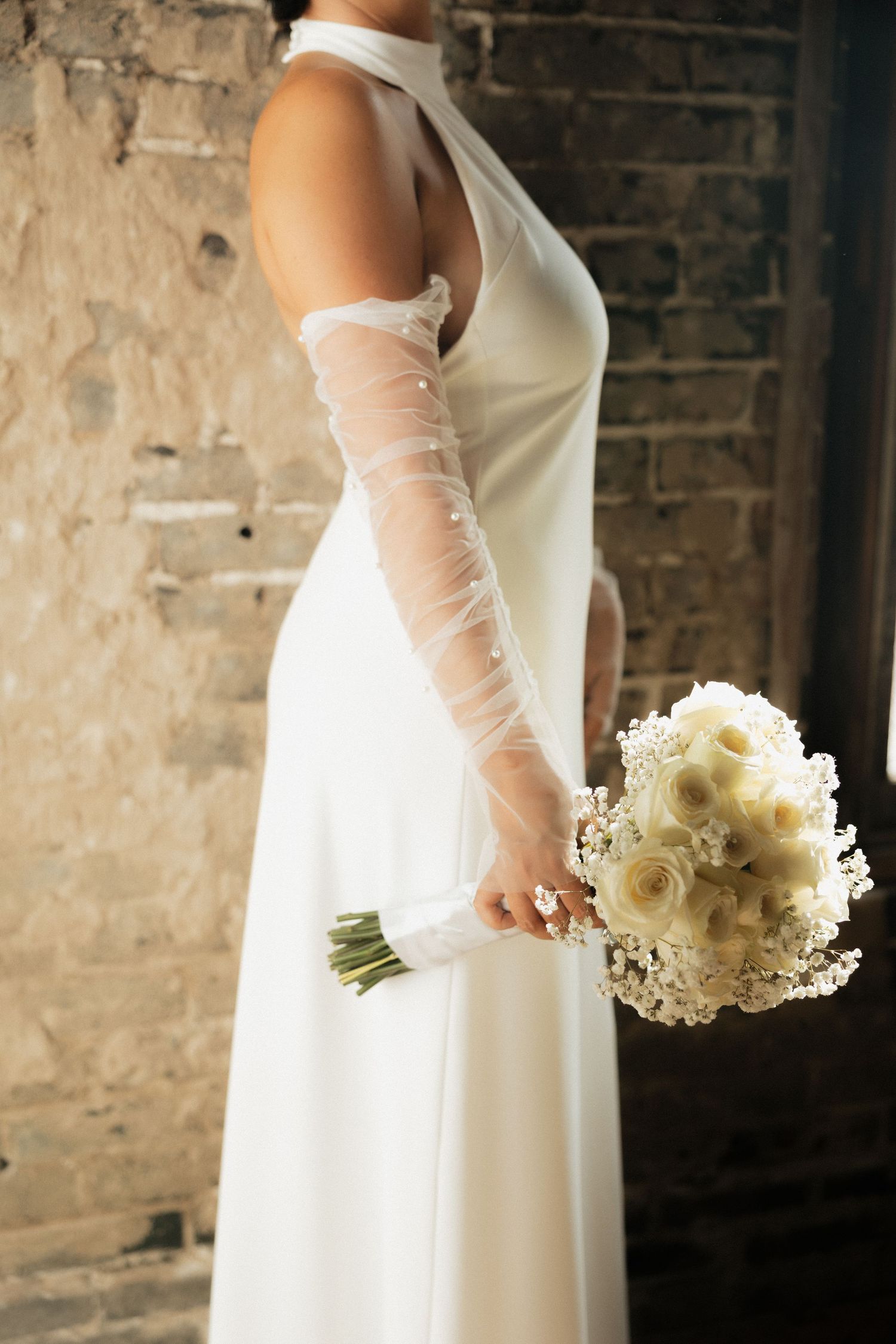 Elegant white wedding dress with long sheer gloves and a delicate white bouquet against a rustic stone wall background.
