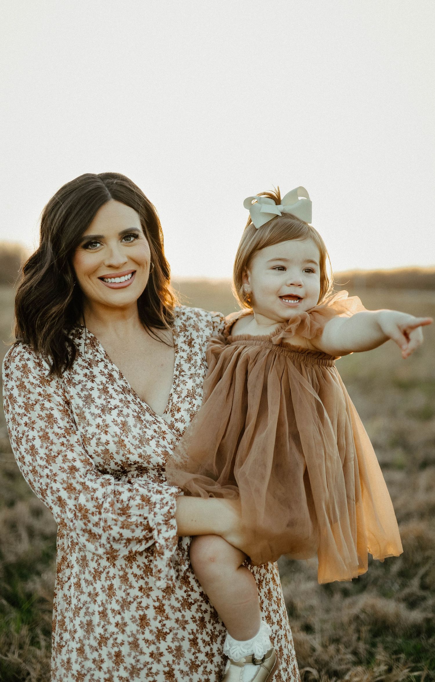 A woman in a floral dress holds a smiling toddler wearing a brown dress and bow in a sunlit field at golden hour.