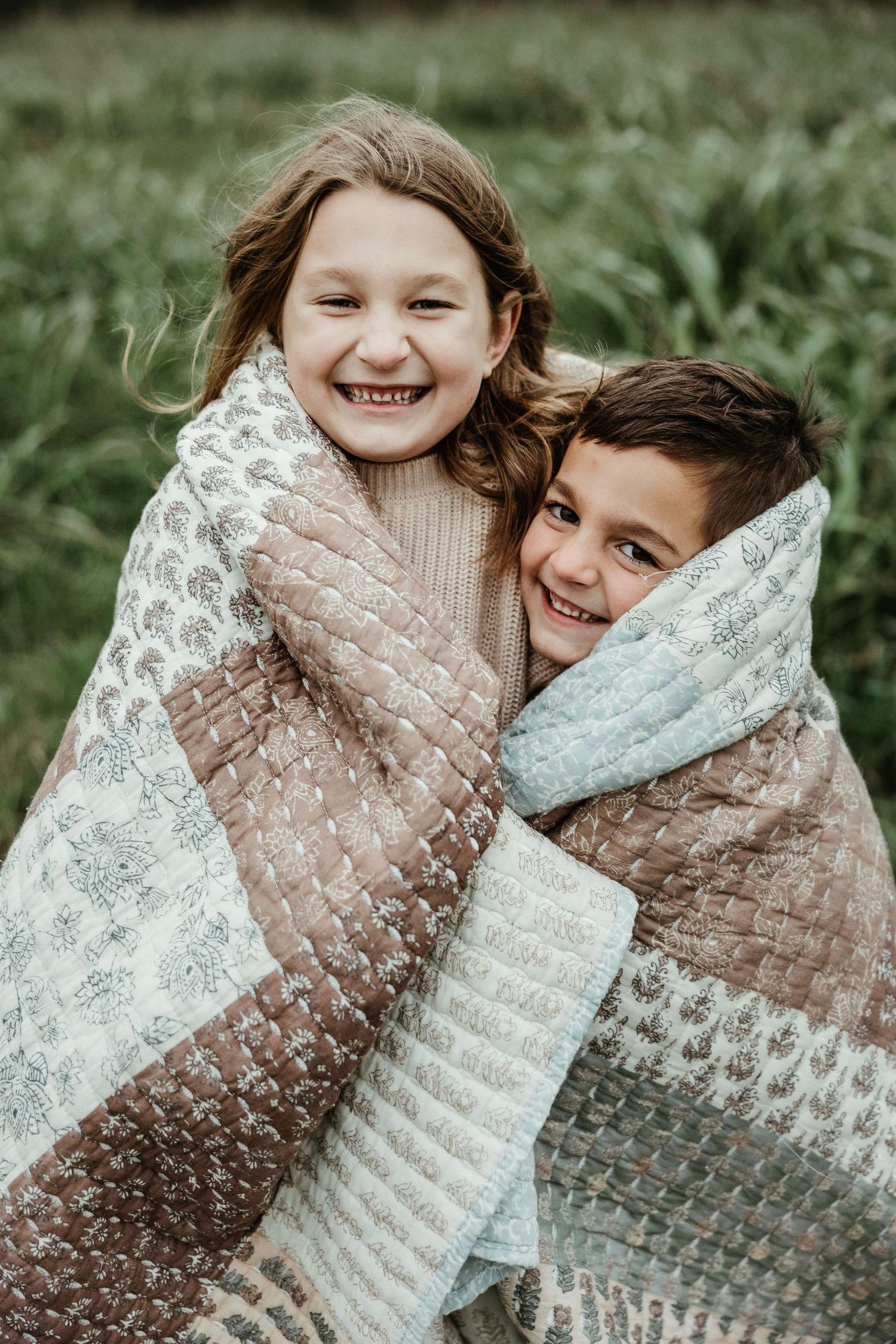 Two children wrapped together in a patterned quilt smile joyfully outdoors against a green nature background.