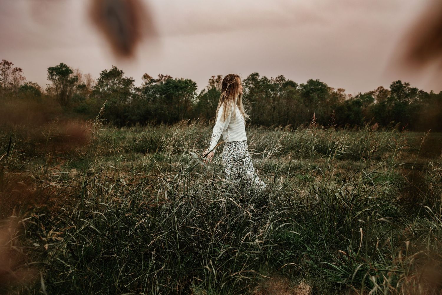 A dreamy moody figure in a white dress stands in a tall grassy field at dusk.