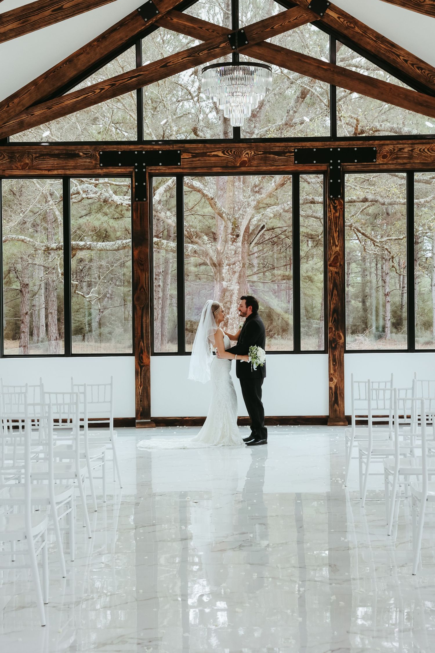 A couple embraces in a modern rustic wedding venue with floor-to-ceiling windows, exposed wooden beams and a crystal chandelier.
