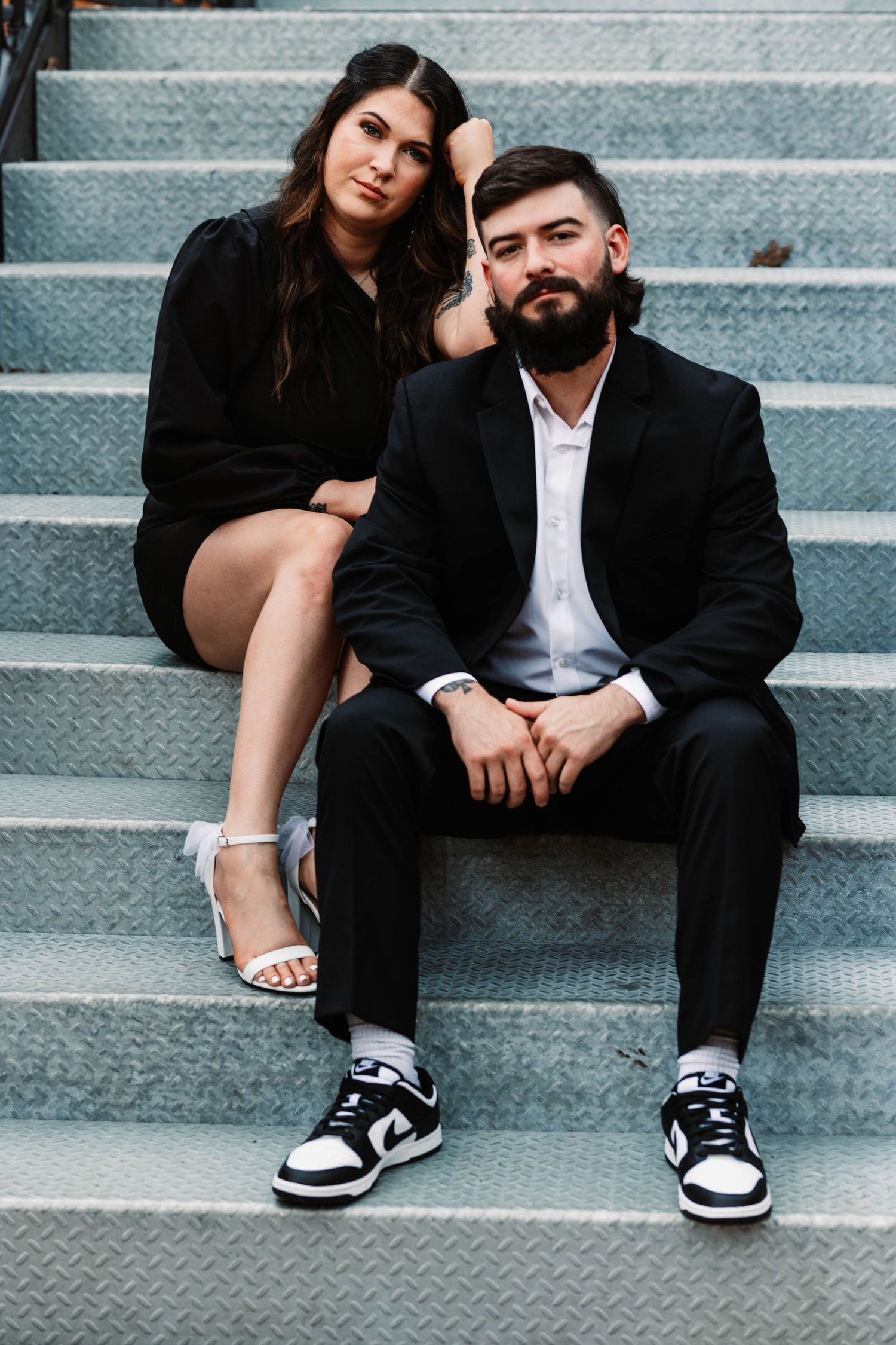 Couple sits casually on concrete steps, wearing contrasting outfits of black formal wear and casual sneakers.