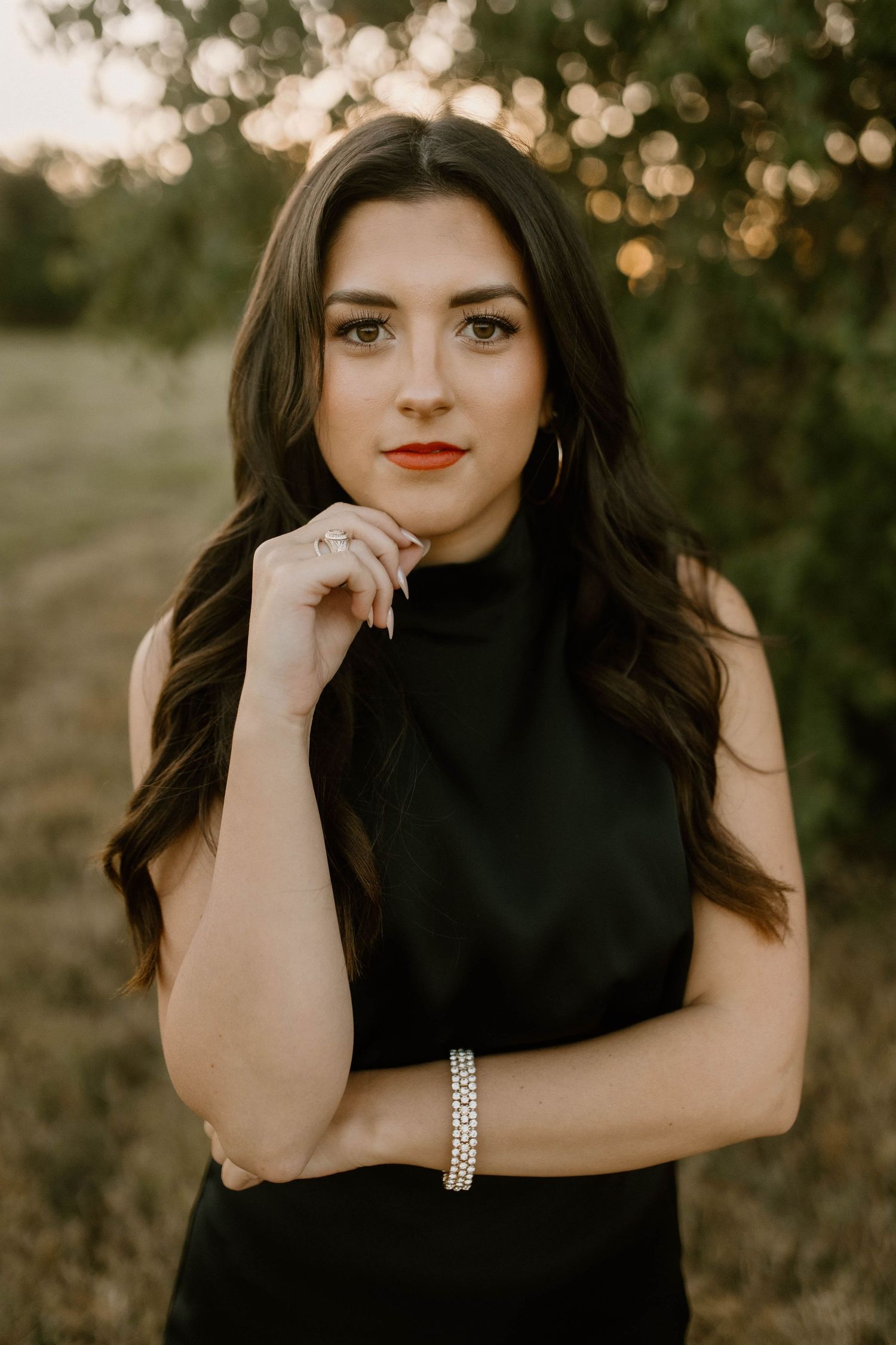 Portrait of woman in black sleeveless top with long dark hair and red lipstick against bokeh sunset background.