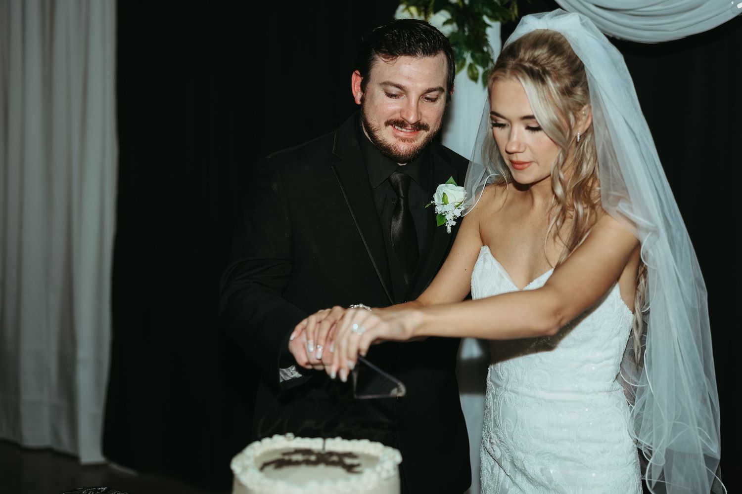 A bride in a white gown and groom in a black suit cut their wedding cake together at their reception.