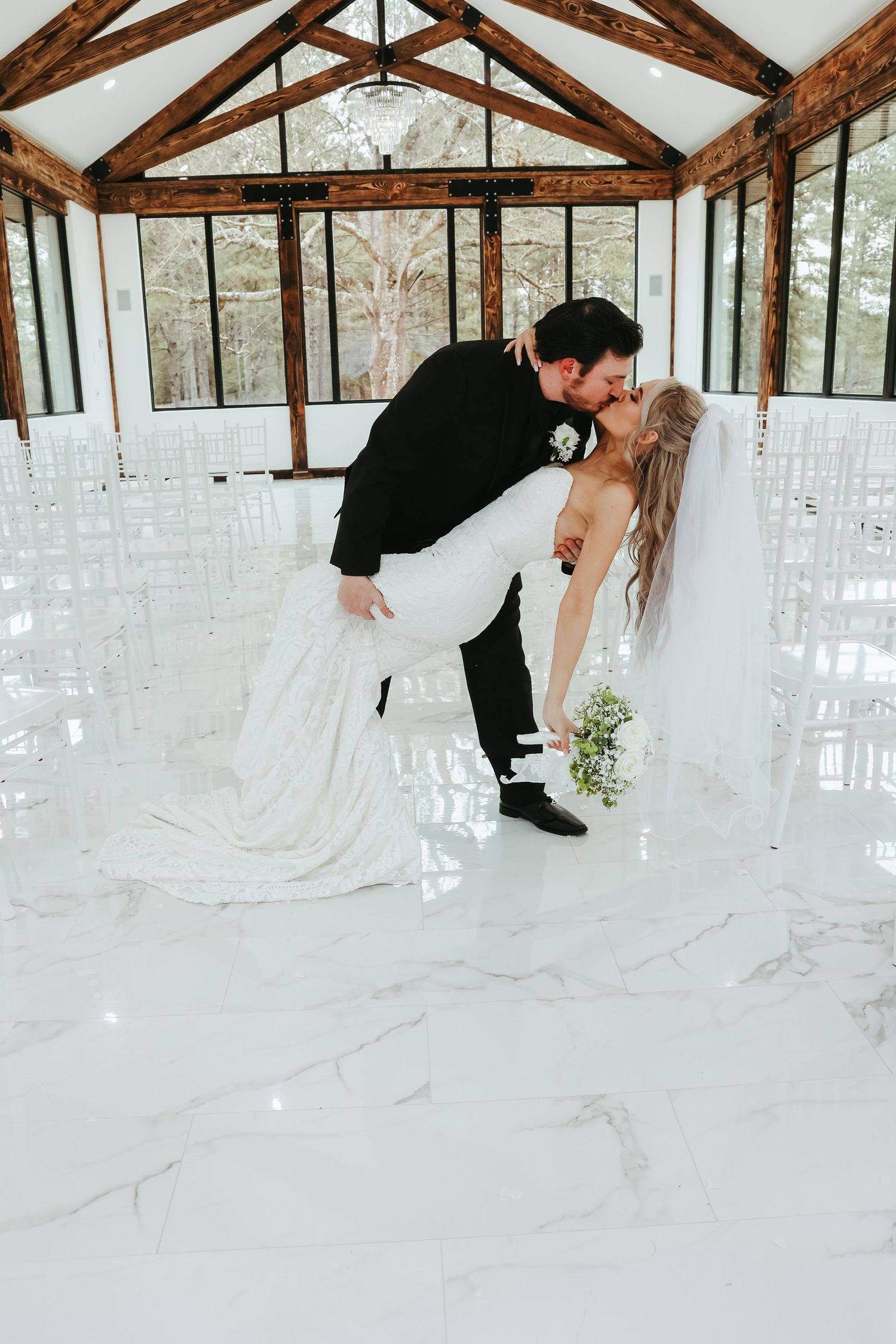 A romantic wedding couple shares a dip kiss in a rustic venue with wooden beams and white marble floors.