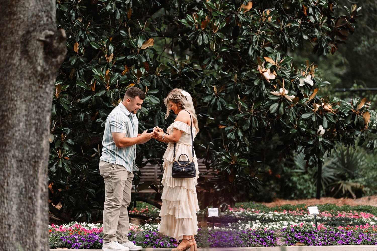A couple shares a romantic moment in a garden with blooming flowers and magnolia trees in the background.