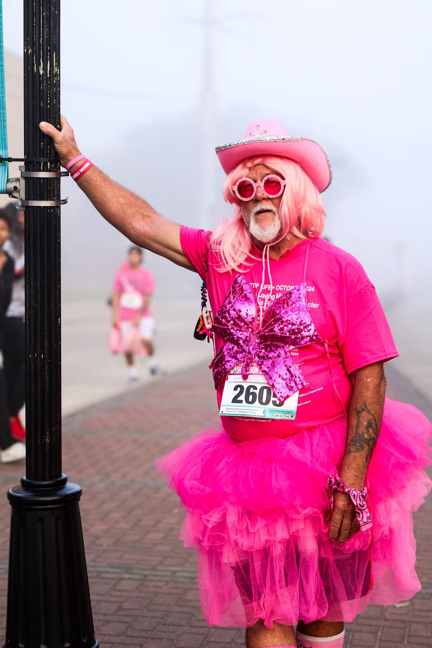 Participant wearing all pink with tutu skirt and sunglasses poses during charity race while holding onto lamp post.