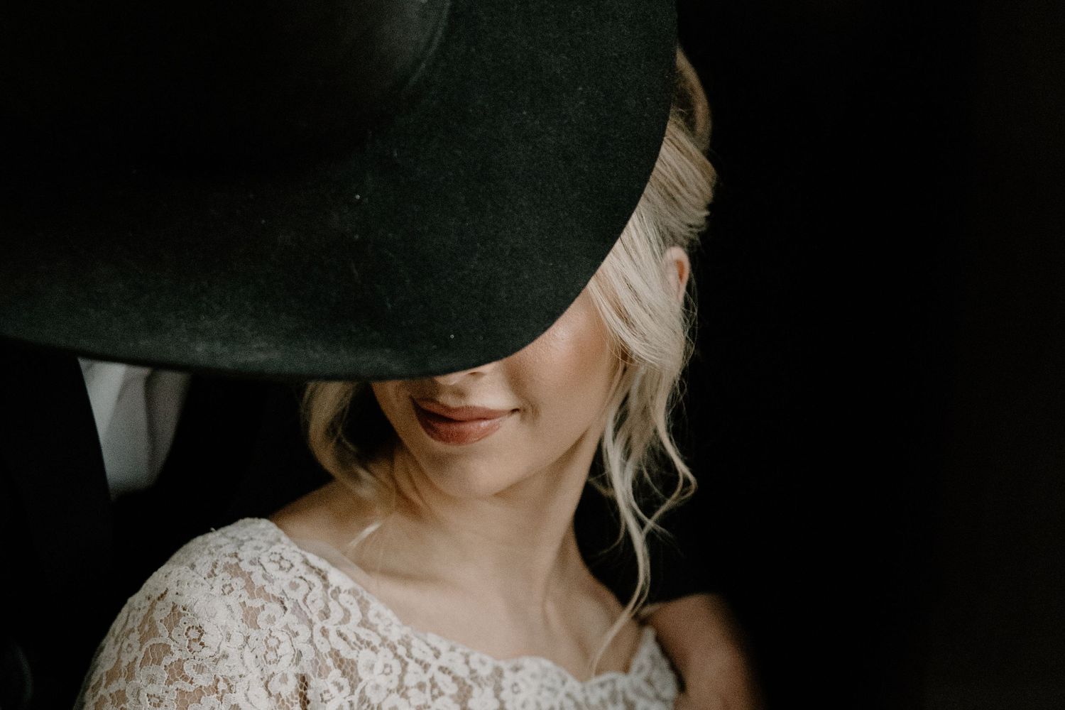 A person in a green hat and lace dress smiles mysteriously in a moody dark portrait.