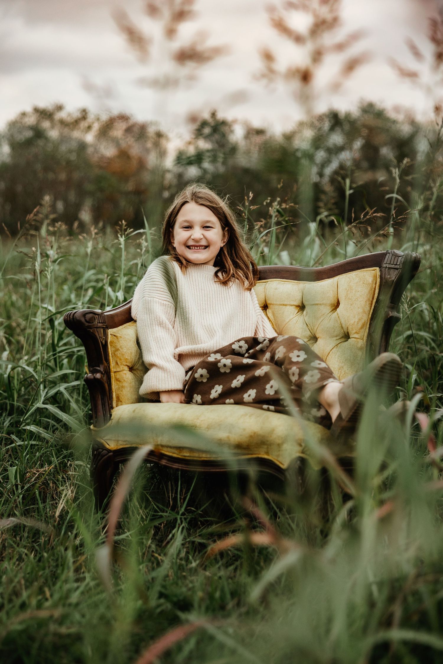 A vintage yellow tufted chair sits in tall grass during golden hour with natural outdoor lighting.