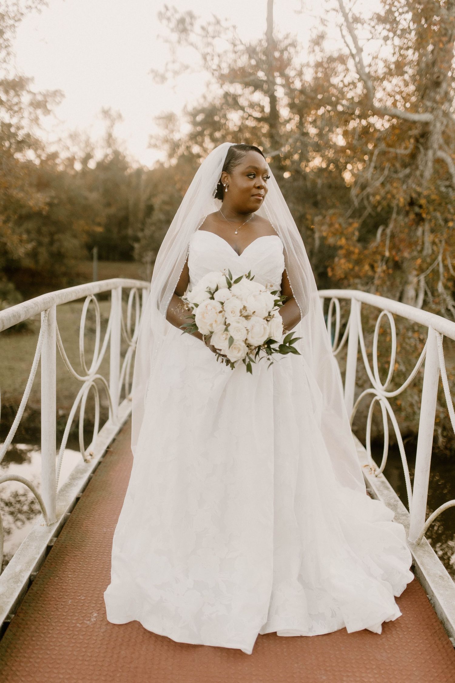 A bride in an elegant white ball gown holds a bouquet of white flowers while standing on a decorative bridge at sunset.