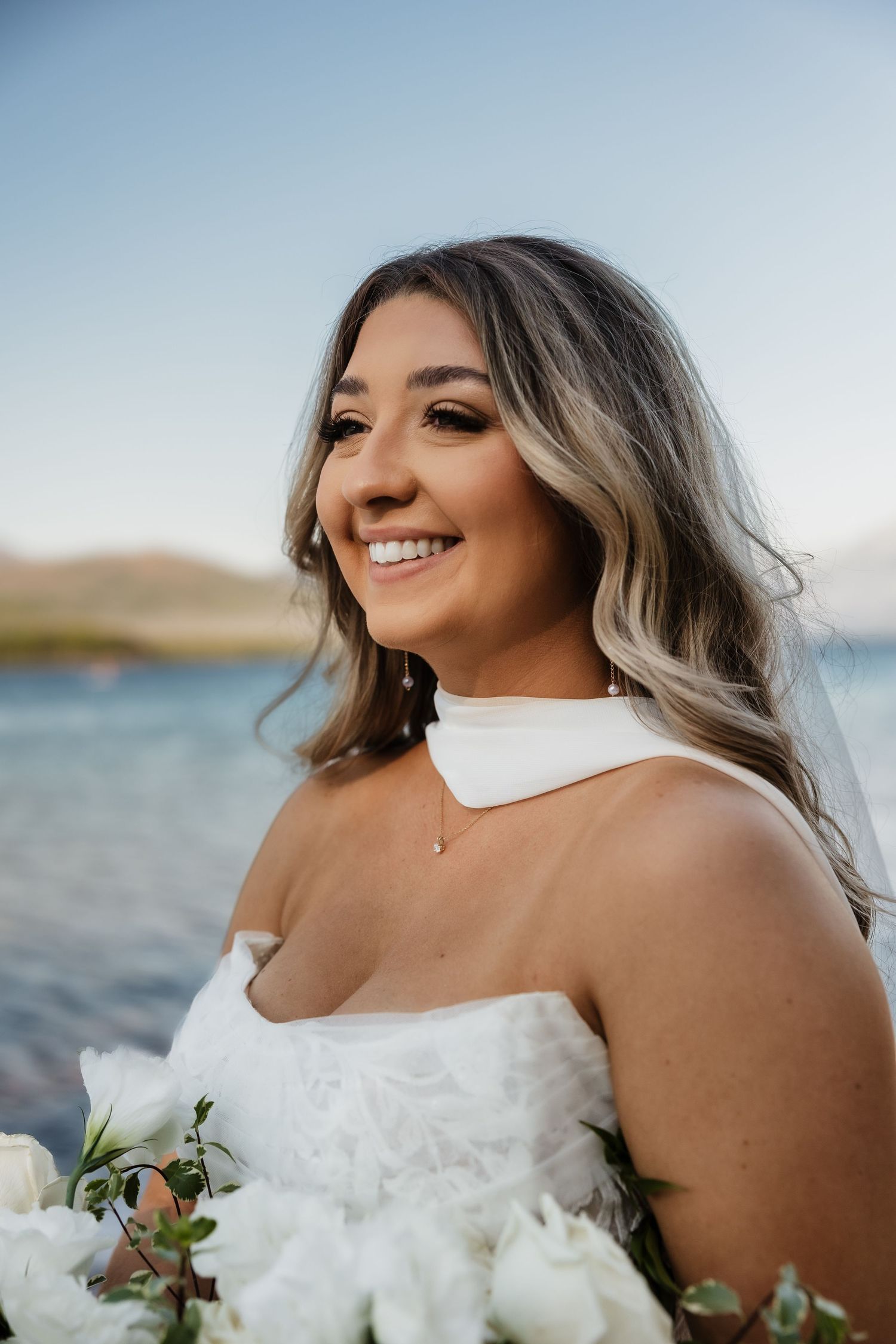 A bride in a white strapless wedding dress smiles while holding a bouquet against a waterfront background.