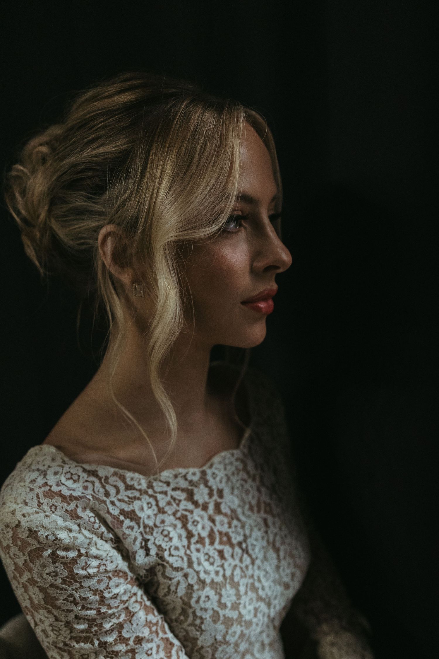 Side profile portrait in dramatic lighting shows blonde woman wearing lace wedding dress against dark background.