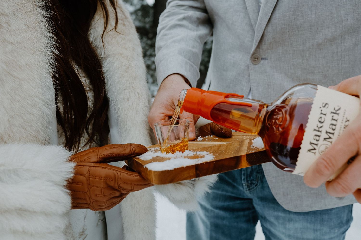 Hands pouring a dark amber-colored whiskey Maker's Mark bourbon into a glass with ice.