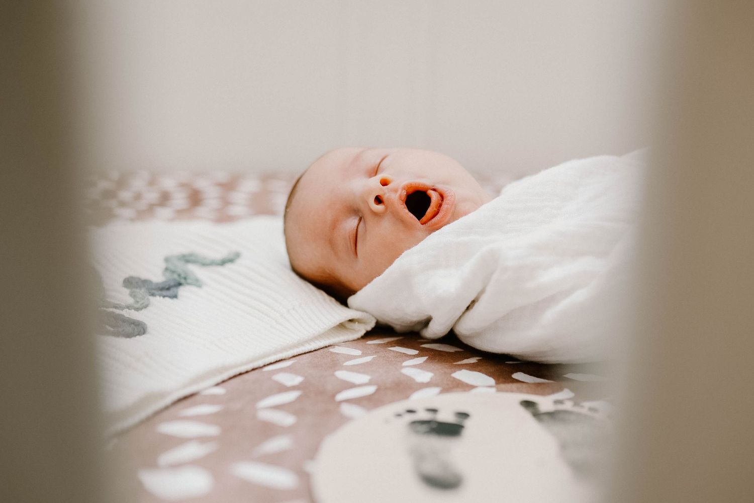 A yawning infant lies on white bedding while wrapped in a cozy blanket.