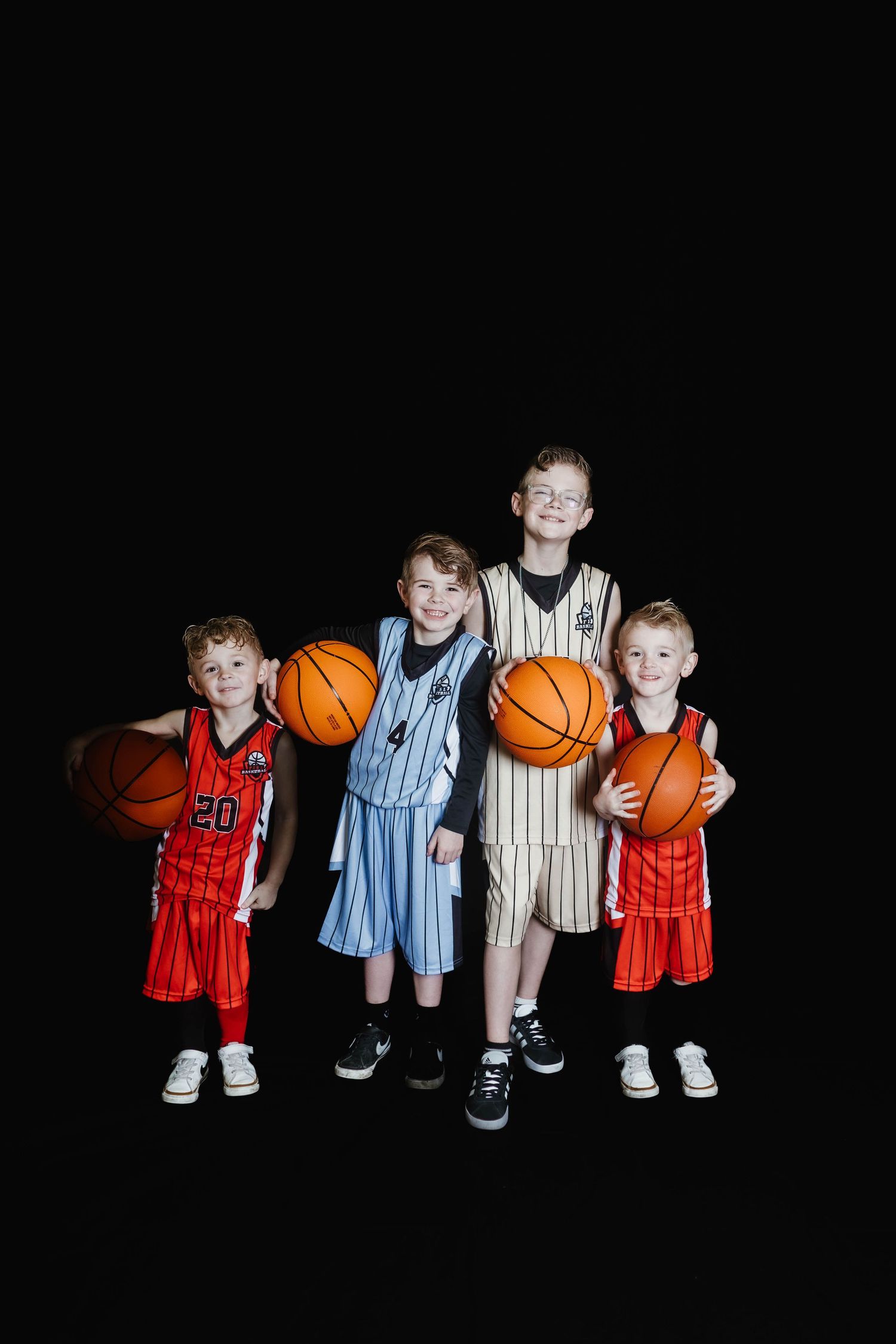 Young basketball players in red and striped uniforms pose together with basketballs against a black background.