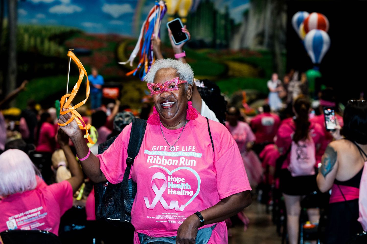 Participant in pink shirt celebrates joyfully at outdoor charity walk event with decorative ribbons and balloons.