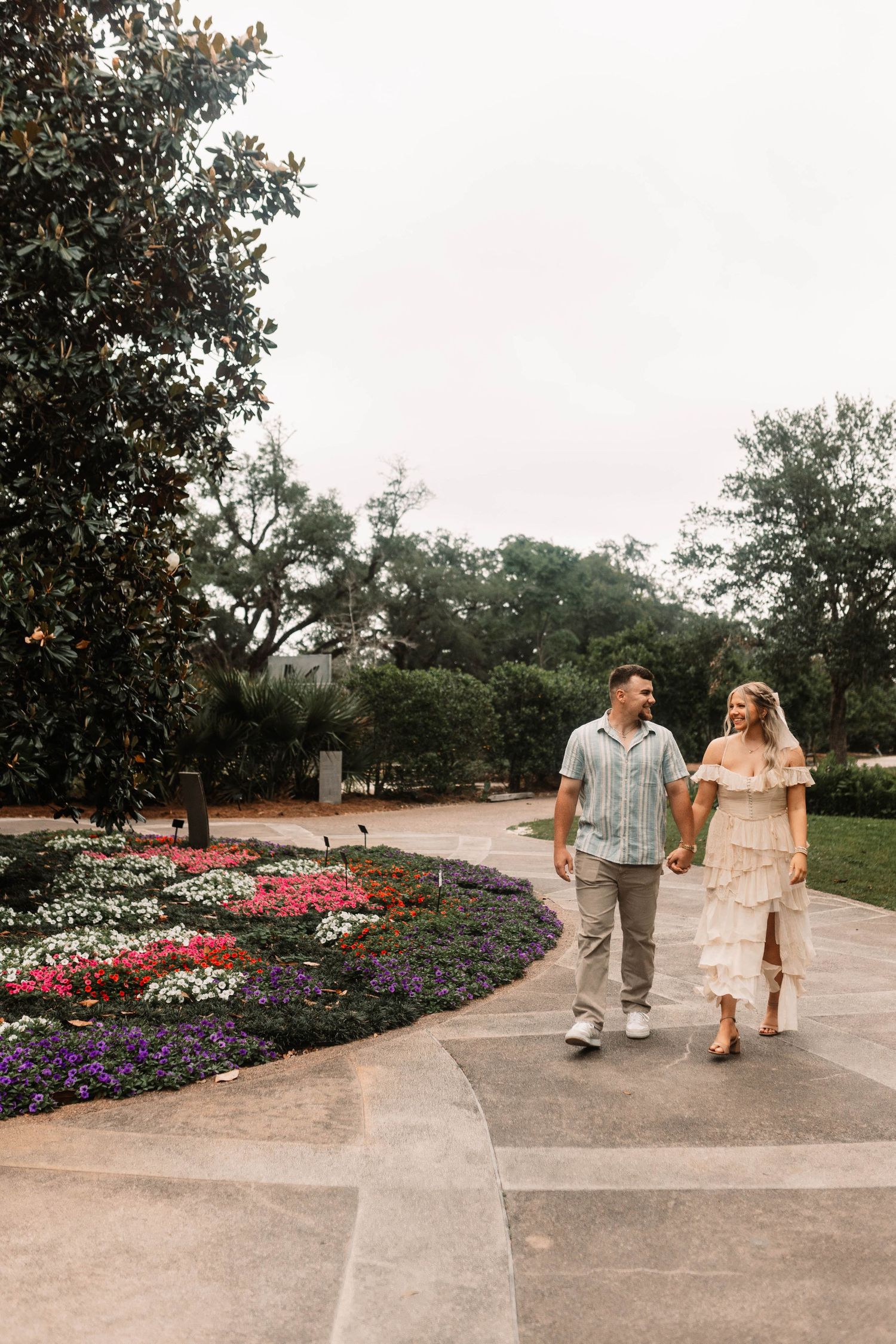 A couple walks hand in hand along a garden path with colorful flower beds and magnolia trees in the background.