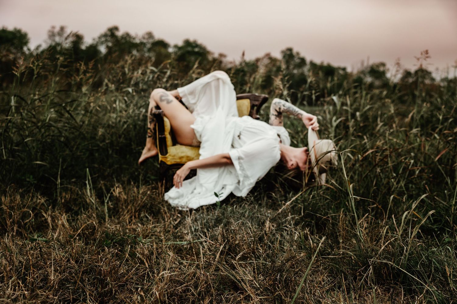 A dreamy ethereal scene of someone in a flowy white dress lying in tall grass at dusk.
