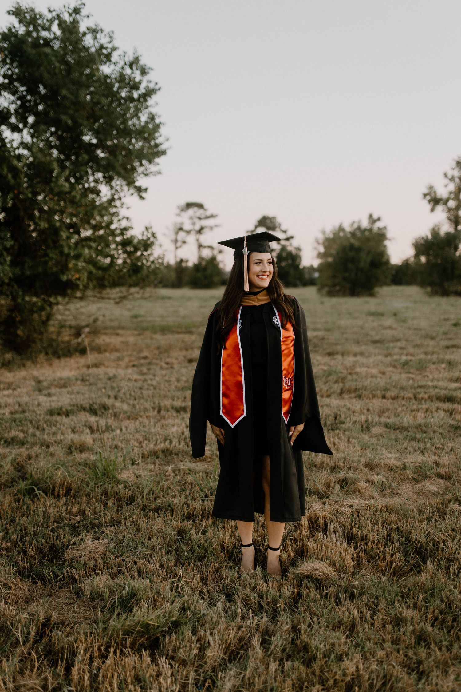 A graduate in black academic regalia with orange and red stoles stands in a grassy field at dusk.