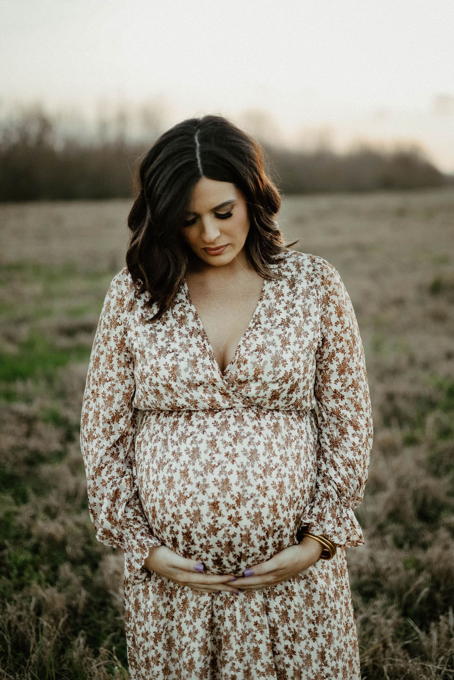 Expectant woman in floral dress stands in field at sunset with hands gently cradling her belly.