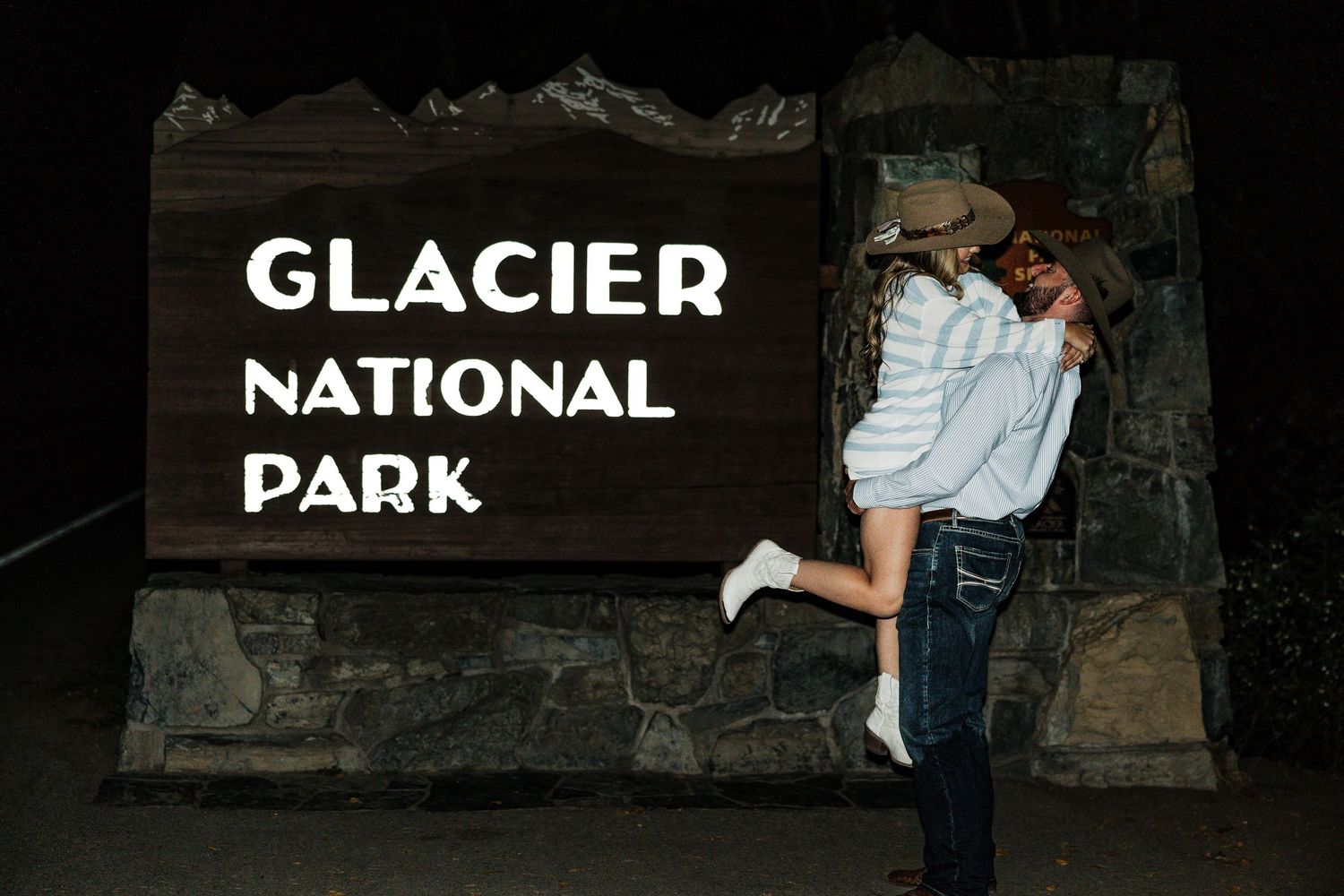 Couple embraces in front of Glacier National Park entrance sign at night.