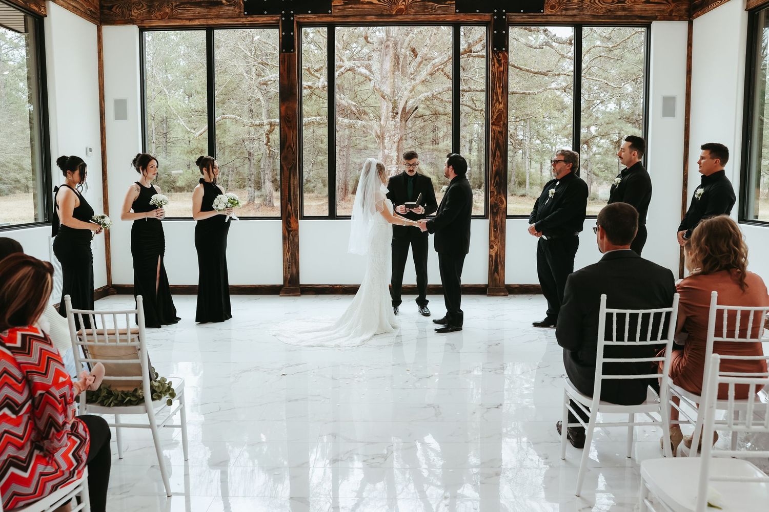 A wedding ceremony takes place in a modern glass-walled venue surrounded by winter forest scenery.