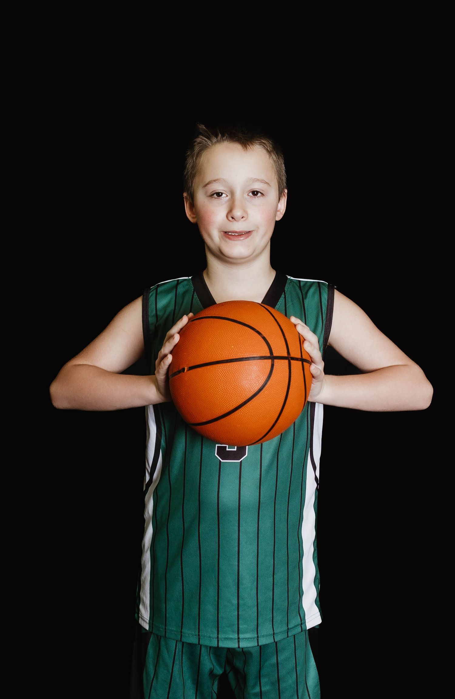 Young athlete in a green and white striped basketball uniform holds a basketball against a dark backdrop.
