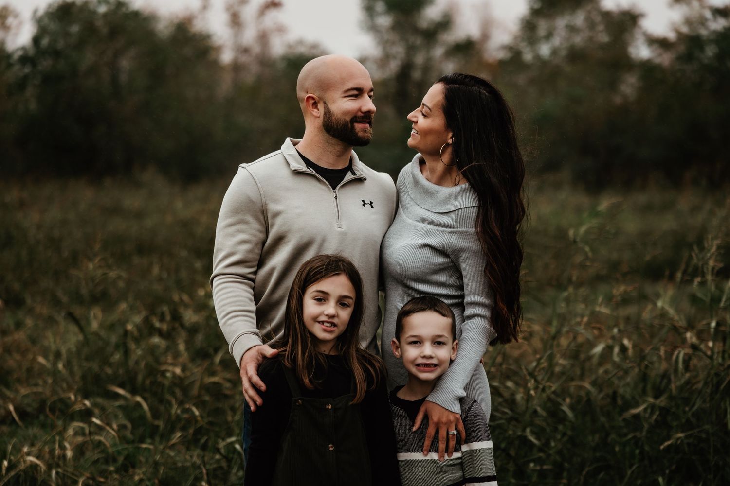 A happy family of four embraces while standing in a tall grass field at dusk.
