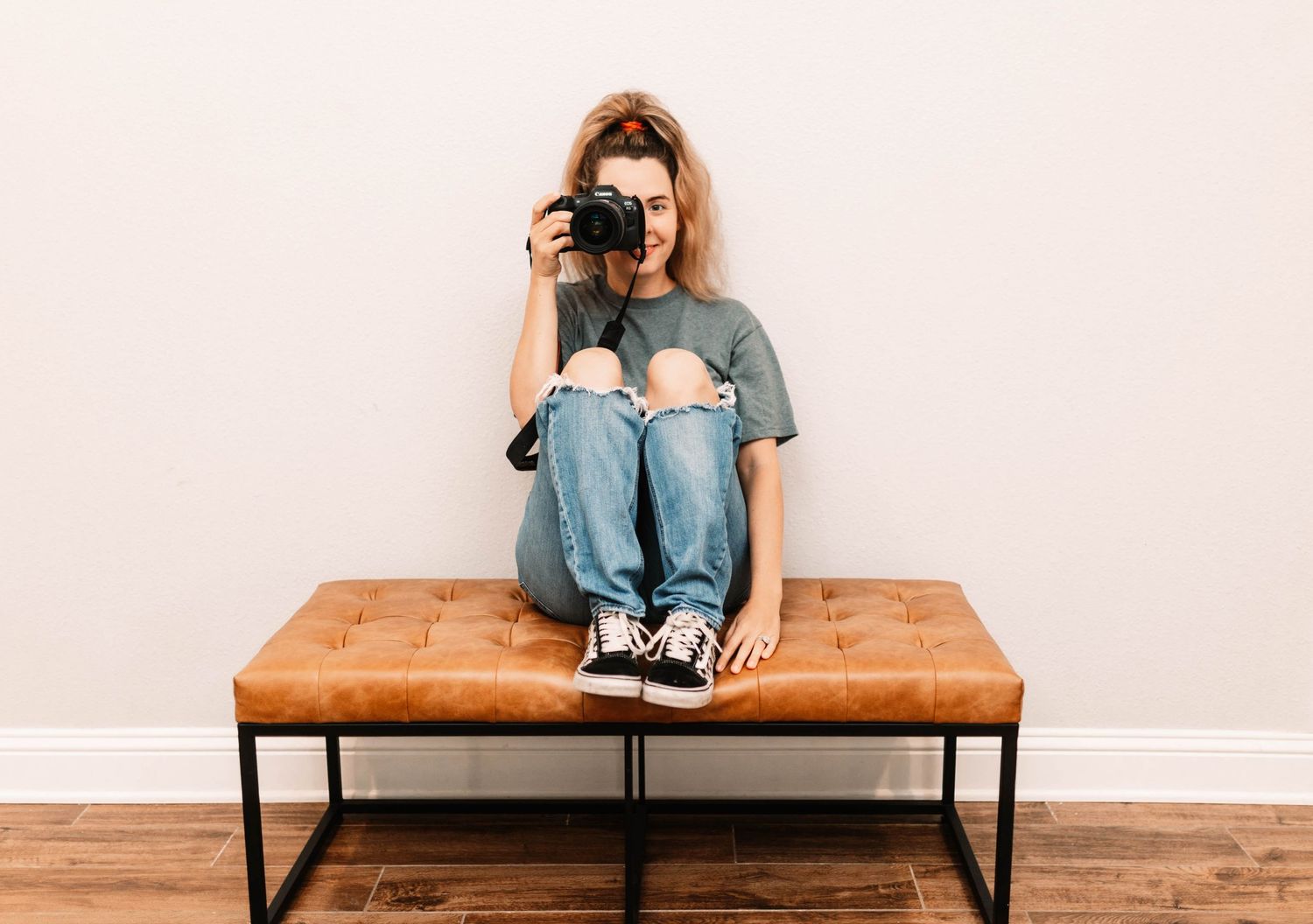 Photographer sitting cross-legged on brown leather bench holds camera in front of plain beige wall.