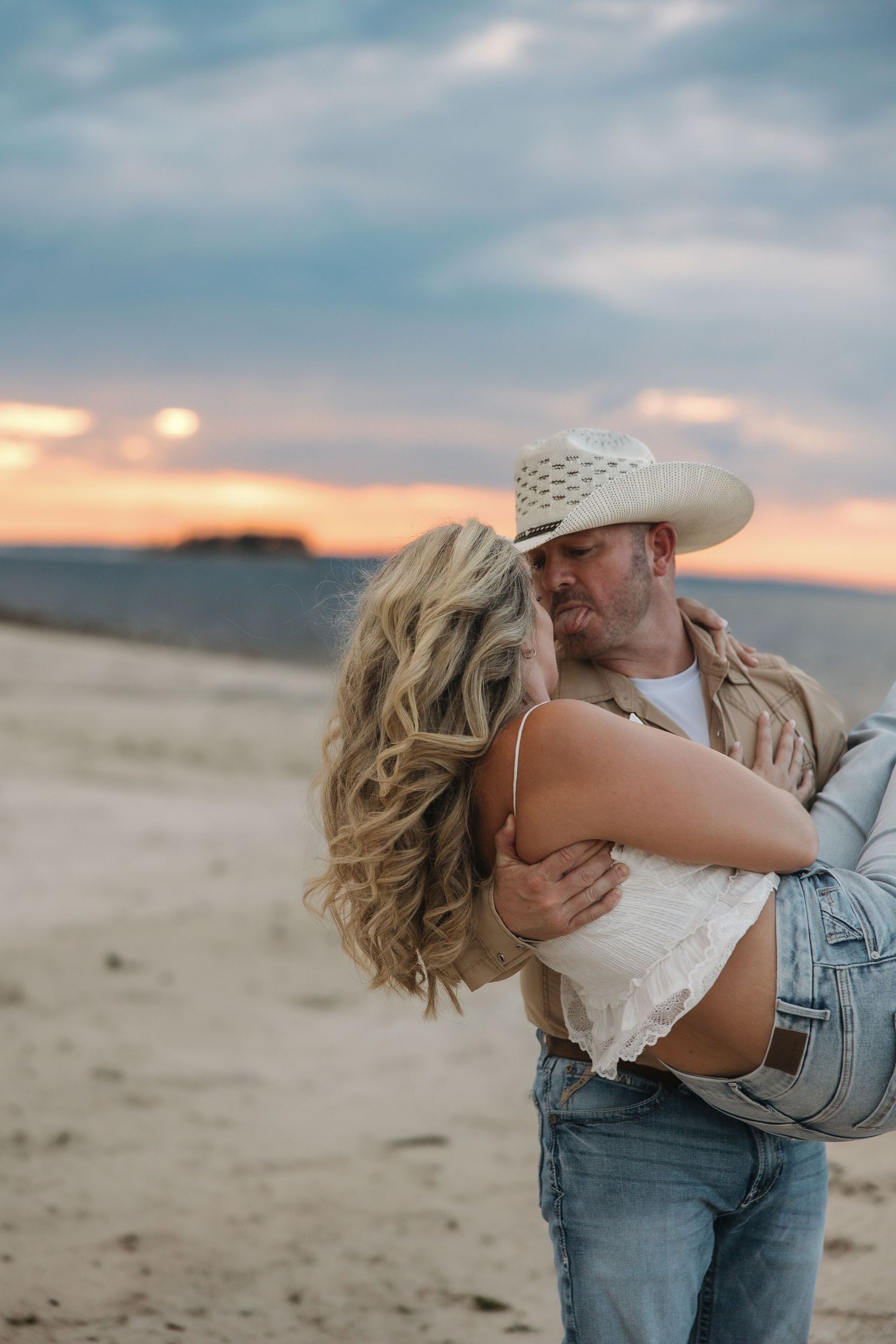 A romantic couple embraces on a sandy beach at sunset with soft pastel clouds in the background.