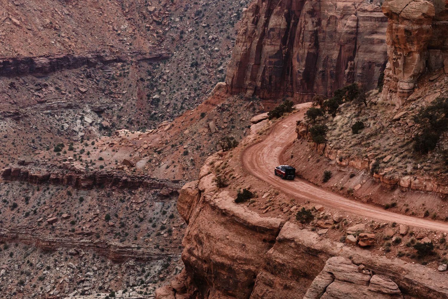 Winding dirt road curves along steep red rock canyon cliffs in a remote desert landscape.