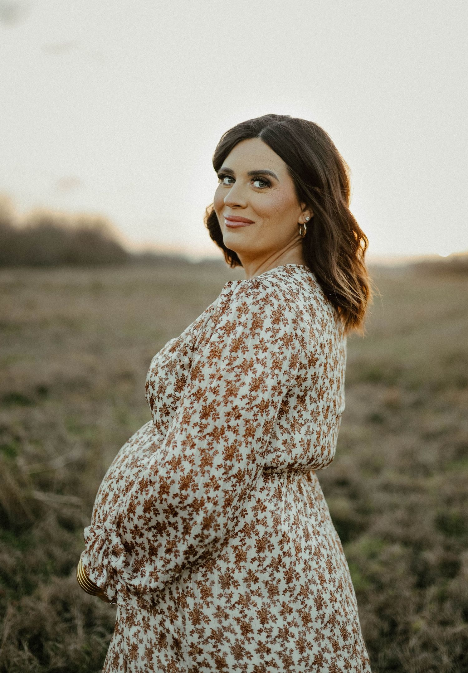 A side profile shot of an expectant mother wearing a floral print dress posing in a field at sunset.
