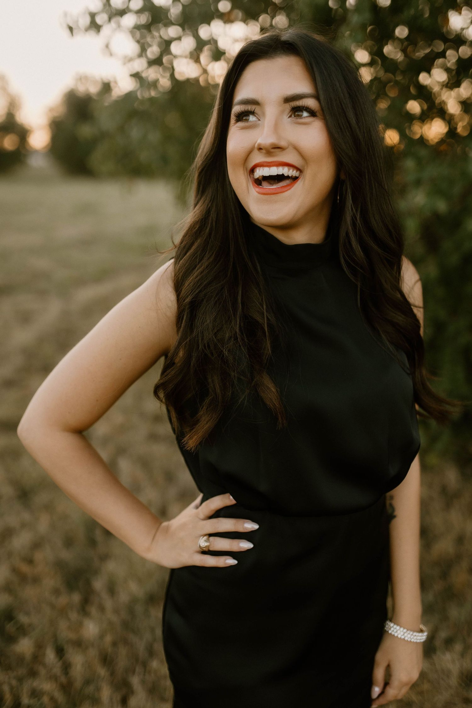 A person wearing a black sleeveless dress poses outdoors during golden hour with a bright smile and bracelet accessory.