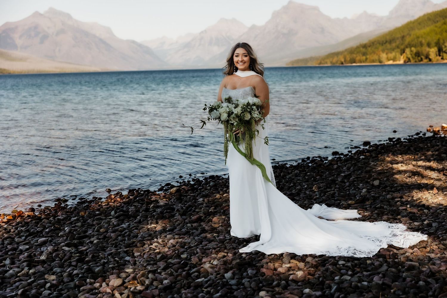 A bride in a flowing white dress holds a green bouquet while standing on a rocky lakeshore with mountains in the background.